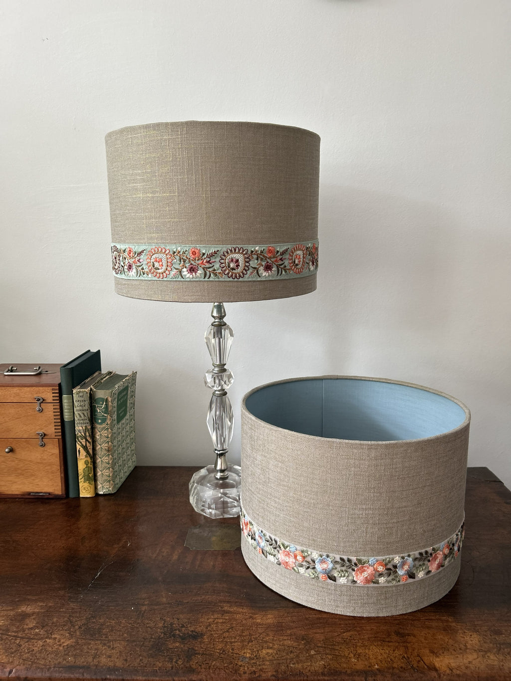 A cylindrical table lamp shade and matching lamp base displayed on a wooden table against a plain wall, alongside books and a small wooden chest. The lampshade and base are made of beige linen-like fabric with a decorative band featuring floral and geometric patterns in muted pink, green, and orange tones.