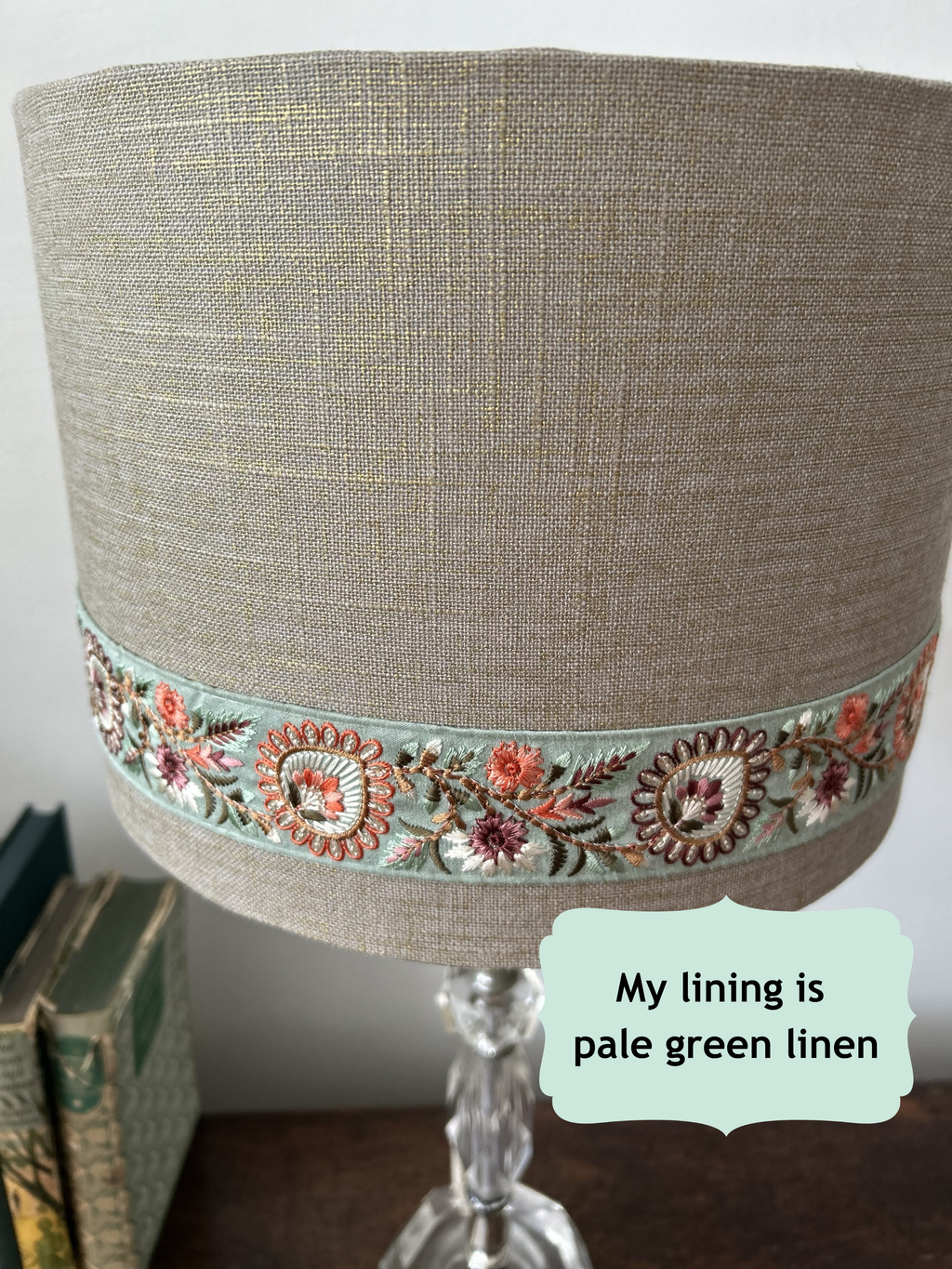 A close-up of a lampshade made from pale gold flecked linen fabric, featuring a decorative band of embroidered floral motifs in pink, orange, and green. A text overlay reads 'My lining is pale green linen'.