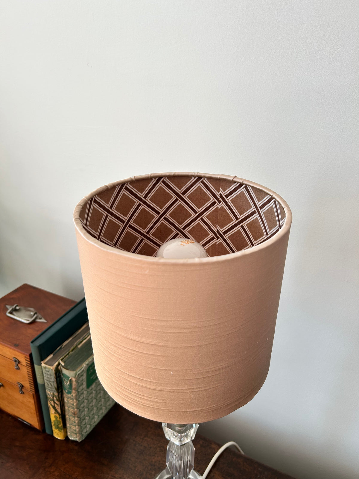 A handmade table lamp shade in a warm peach-beige colour with a brown and white geometric lattice pattern inside, shown on a glass lamp base next to vintage books.