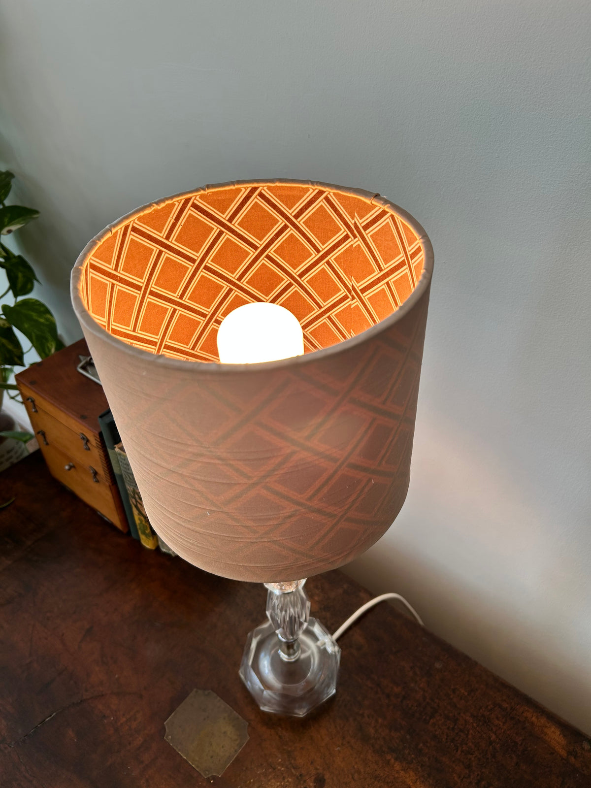 A table lamp with a drum shade featuring a textured beige and coffee geometric diamond pattern, mounted on a clear glass base and shown lit on a wooden table against a light gray wall.