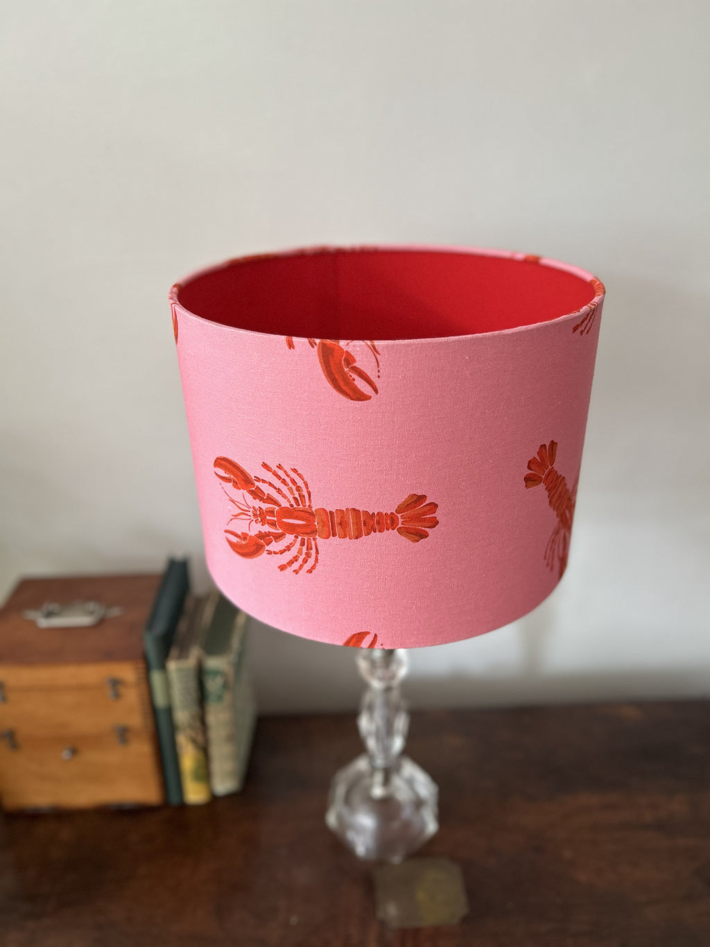 A pink table lamp shade with a red lobster pattern, mounted on a clear glass pedestal base on a wooden table.