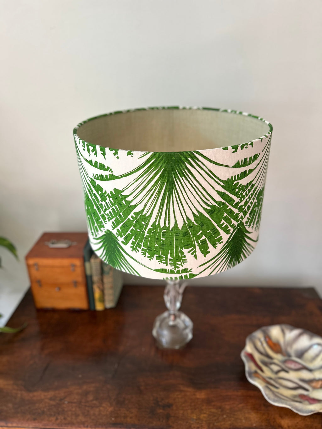 A table lamp shade with a green palm leaf pattern on a cream background, mounted on a clear glass stem base and set on a wooden table.