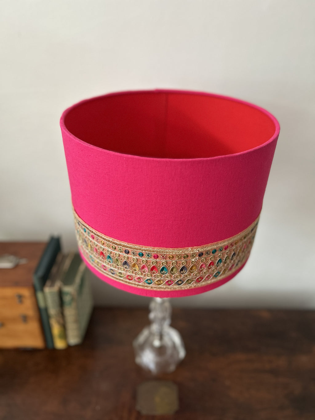 A bright pink cylindrical lampshade with an ornate gold embroidered band featuring colorful rhinestones, displayed on a clear glass pedestal stand on a wooden surface.