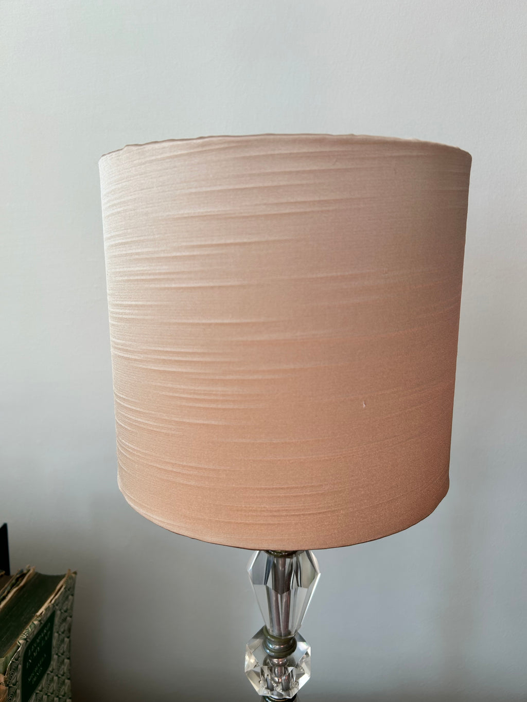 A cylindrical fabric table lampshade in a dusty blush pink color, shown on a table lamp with a faceted clear crystal base against a plain light gray wall.