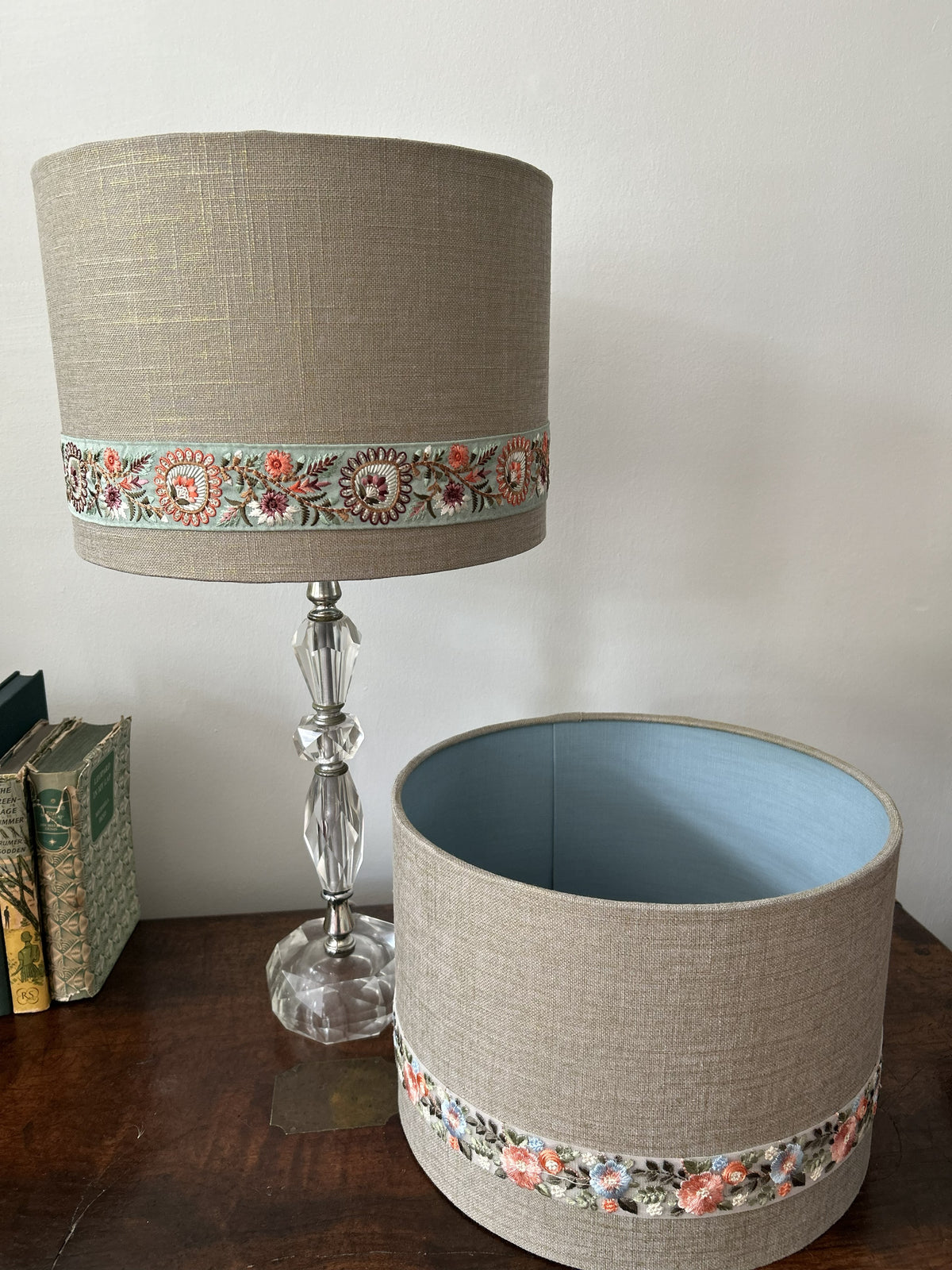 A cylindrical fabric lampshade in a warm beige linen-like material with a decorative floral band around the lower portion, shown next to a matching lamp base on a wooden table against a plain wall.