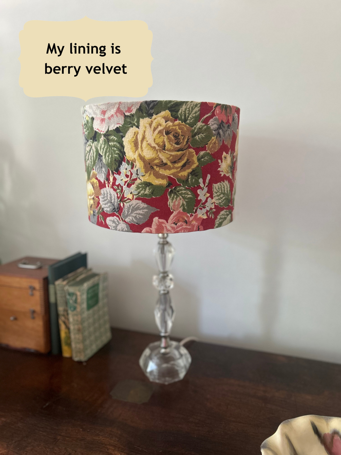 A table lamp with a cylindrical drum shade featuring a vintage floral print in red, yellow, green, and pink, mounted on a clear glass pedestal base. The lamp sits on a dark wooden table next to some books against a plain white wall, with a text bubble reading 'My lining is berry velvet'.
