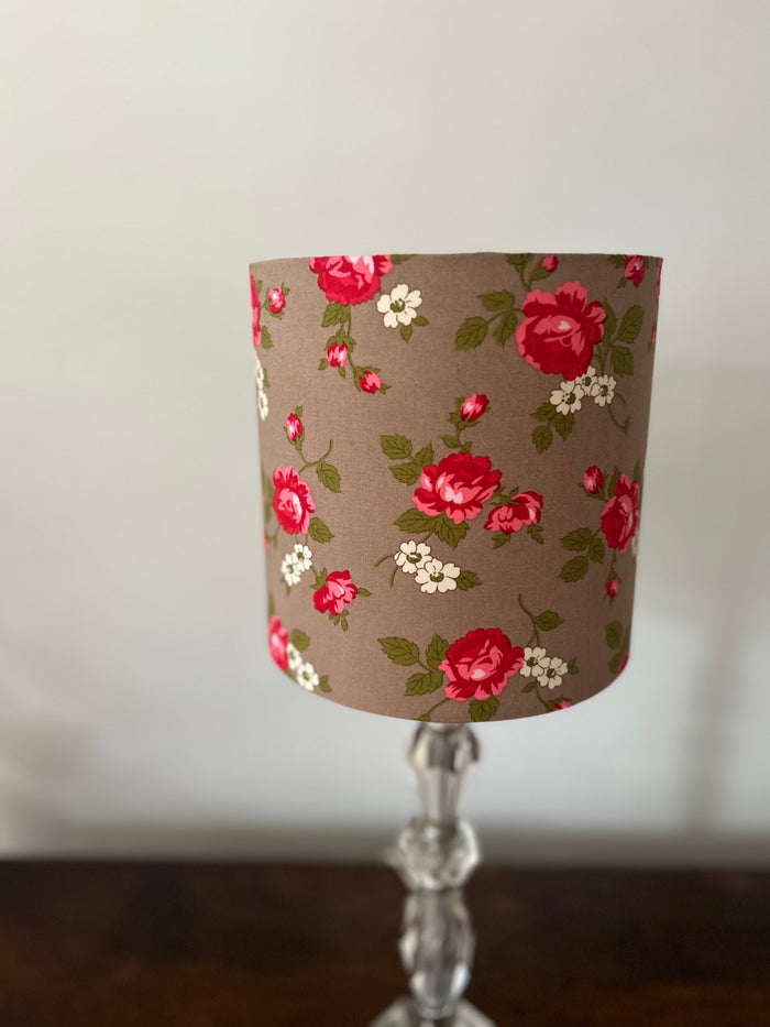 A fabric lampshade with a red rose and white floral pattern on a brown background, the fabric is called 'scattered rosebuds', displayed on a clear glass candlestick base. The shade has a vintage cottage core feel about it.