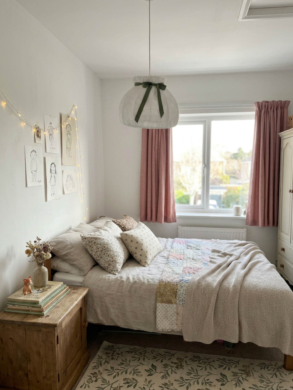 A cozy bedroom scene featuring a bed dressed in beige and multicolor patchwork bedding, styled with pink curtains, a wooden bedside table, and a hanging pendant light.