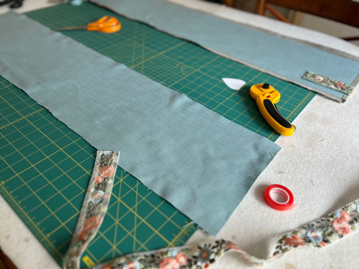 A photo of a drum lampshade in production. A long strip of dusty blue-grey linen fabric laid out on a green cutting mat, with sewing tools including a rotary cutter, scissors, and floral embroidered ribbon visible around it.