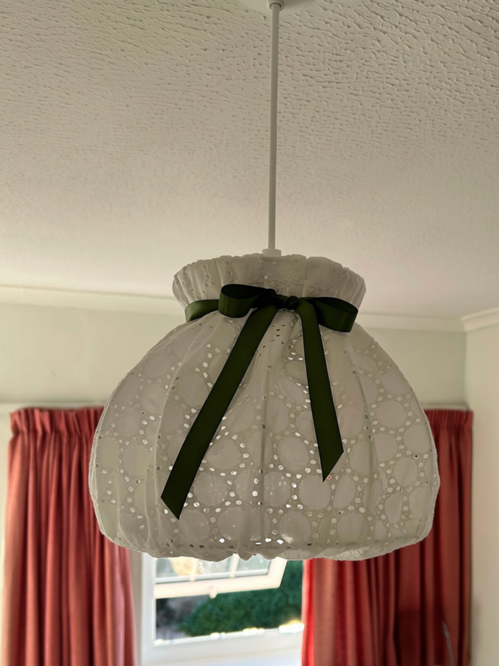 White broderie anglaise lampshade with a green satin bow, suspended from a ceiling fitting in front of pink curtains.