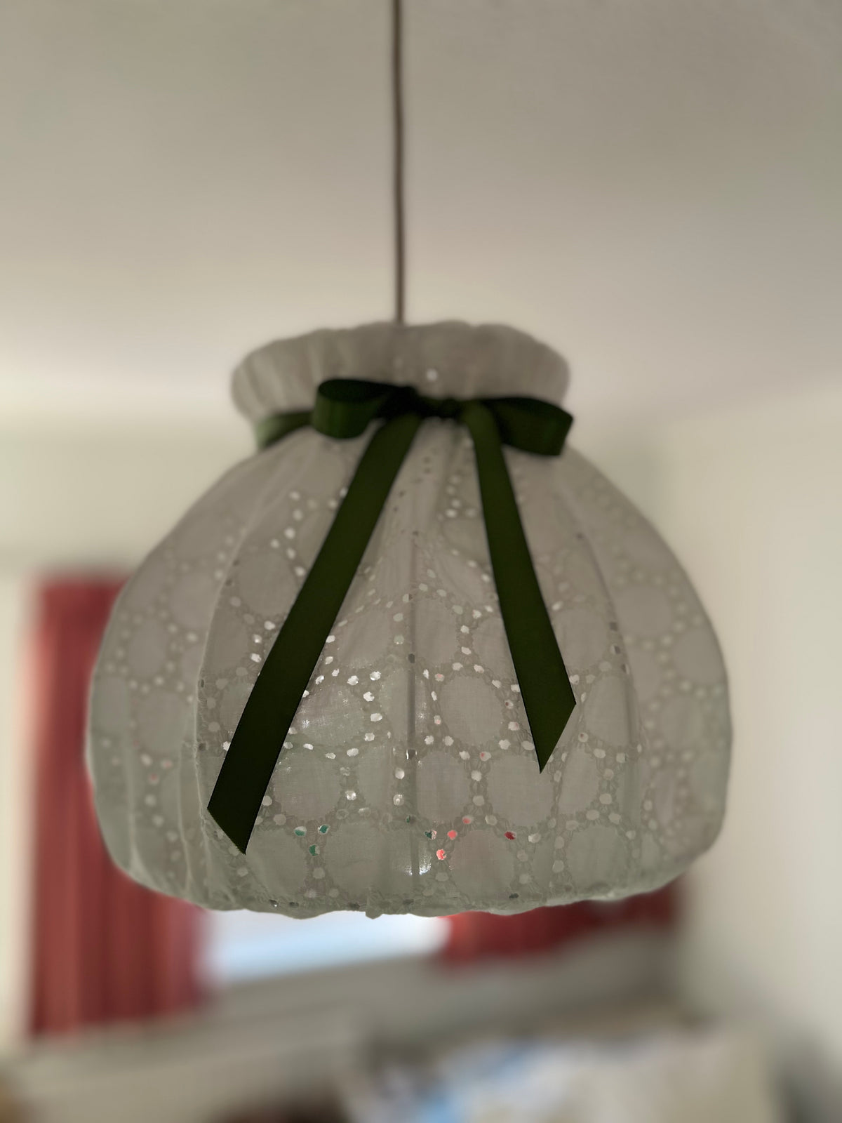 Unlit white broderie anglaise pendant shade, showing its curved panels, scalloped lower edge and sage-green bow.