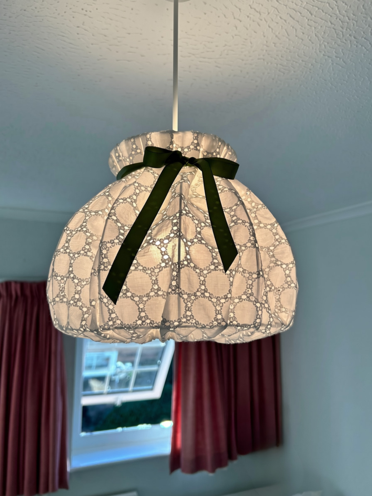 Lit white broderie anglaise pendant lampshade, with light revealing its embroidered eyelet pattern beneath a green satin bow.