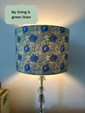 A table lamp with a cylindrical drum shade featuring a blue and green floral William Morris style damask pattern, lit to cast a warm glow against a blue wall. A text bubble in the top left reads 'My lining is green linen'.