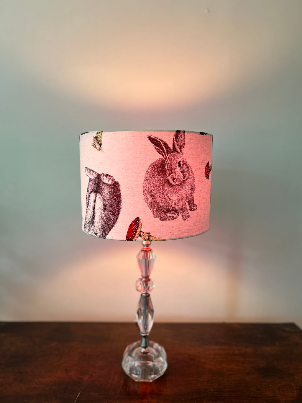 Lit rabbit-and-toadstool drum lampshade on a faceted glass base, casting warm light above and below the shade.