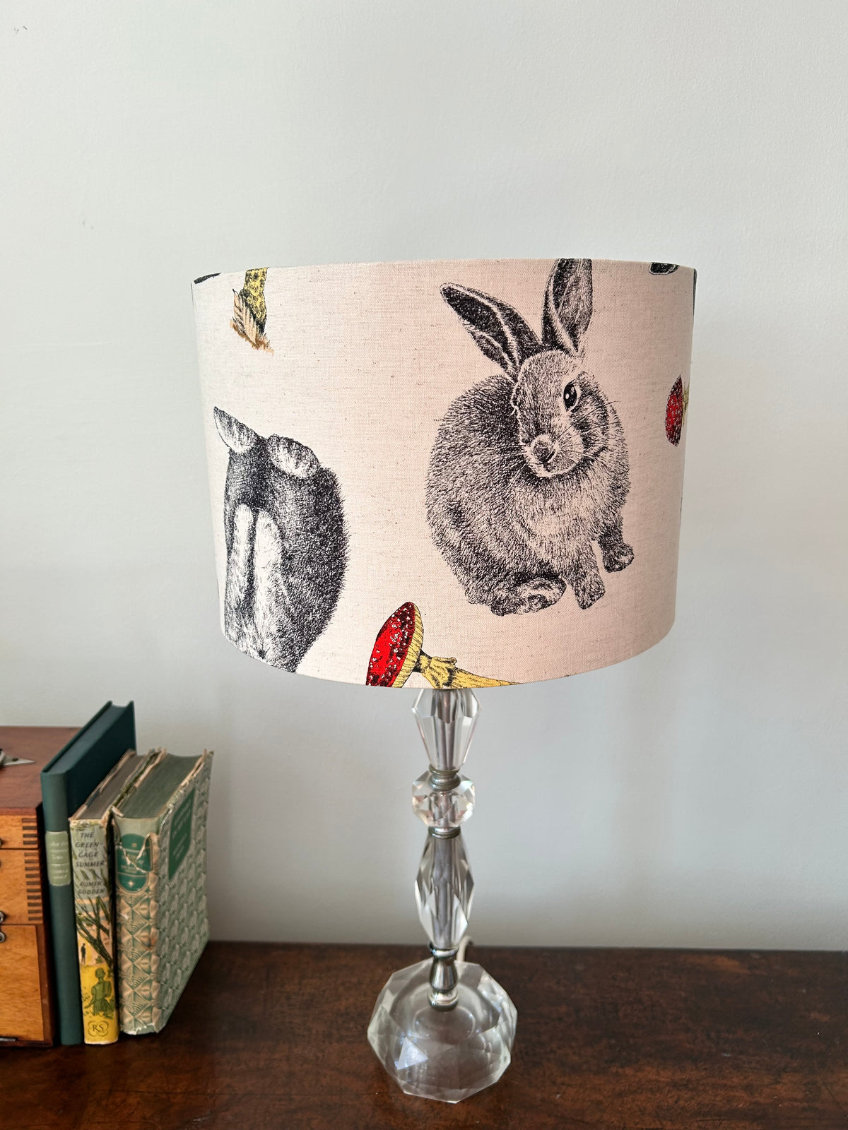 Unlit rabbit-and-toadstool drum lampshade on a clear glass base beside vintage books, with grey rabbits on cream fabric.