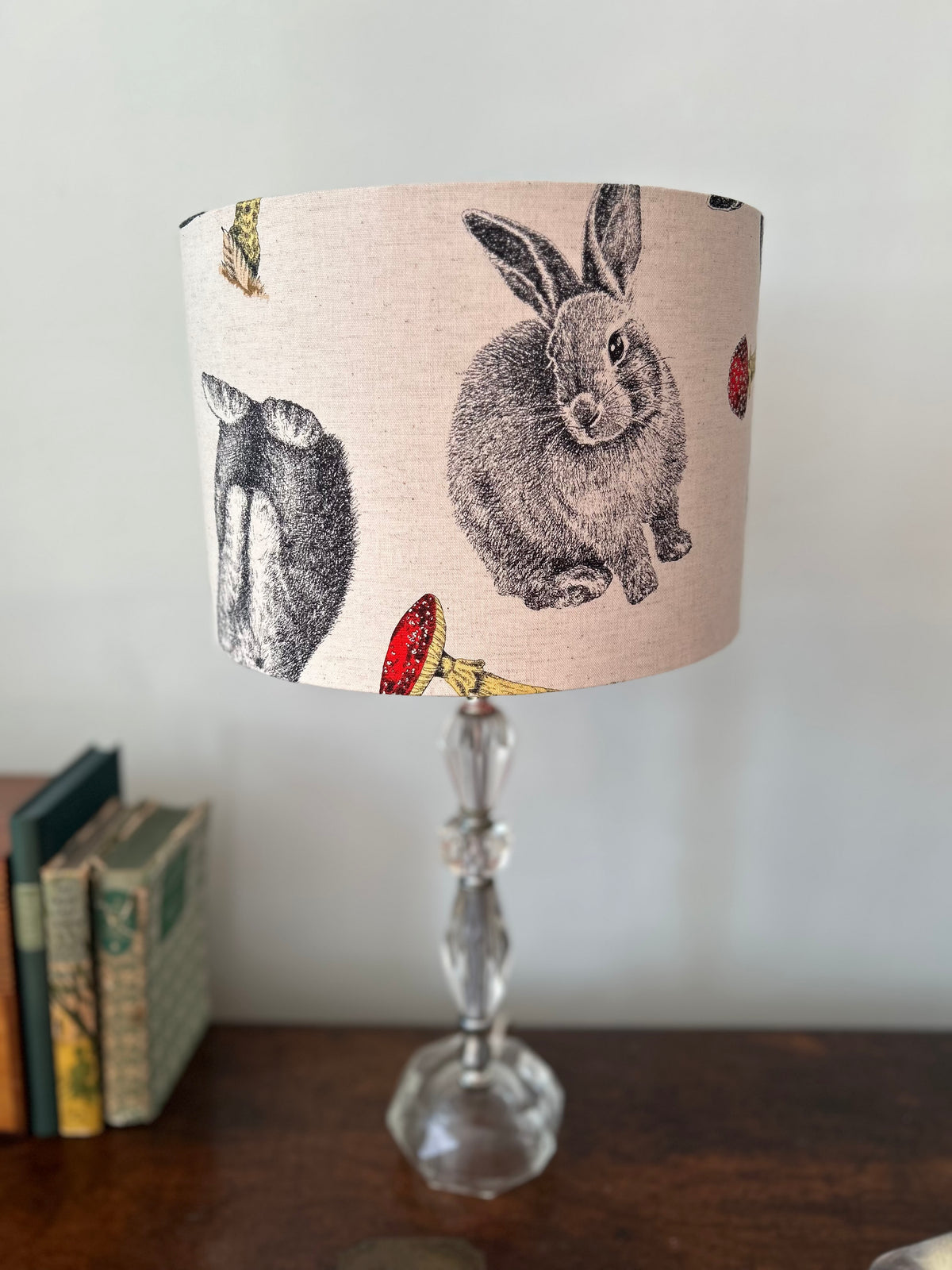 Close view of an unlit woodland drum lampshade, showing finely drawn grey rabbits and red toadstools on cream fabric.