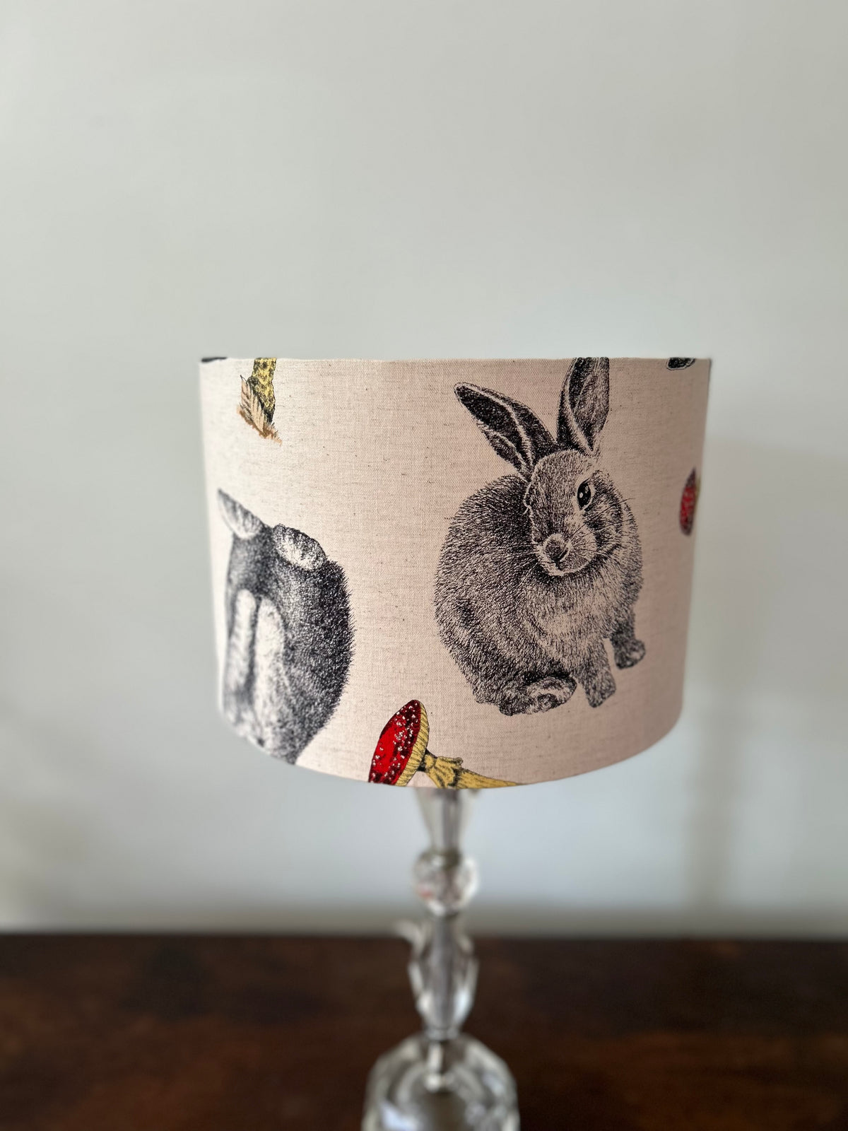 A table lamp with a beige fabric drum shade featuring black and gray rabbit illustrations and small red and yellow accents, mounted on a clear glass stem base.