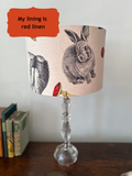 A table lamp with a beige fabric shade featuring black rabbit illustrations and small red toadstools, set on a dark wooden table against a light wall. An orange text box in the top left reads 'My lining is red linen'.