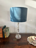 Alt text: Blue velvet drum lampshade on a clear glass table lamp beside books on a wooden sideboard.