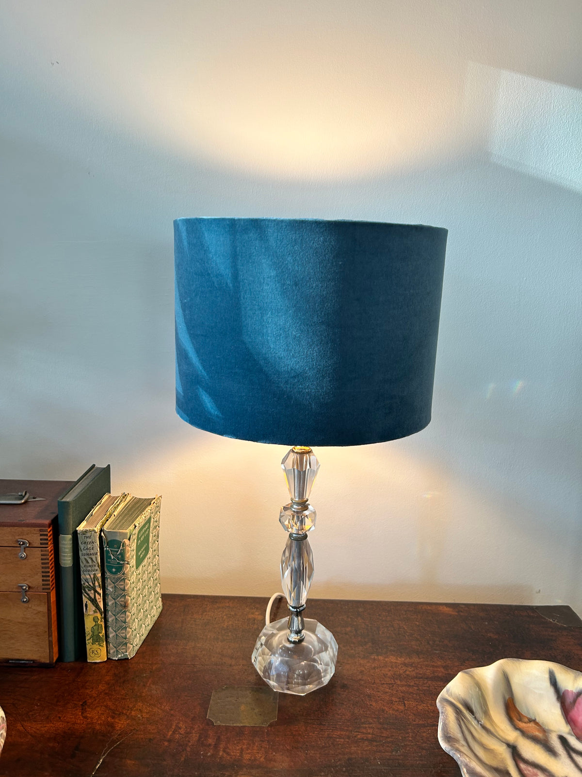 Illuminated blue velvet lampshade on a glass table lamp, casting warm light onto the wall.