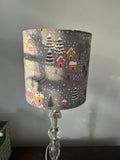 Grey Christmas drum lampshade with raspberry and mustard cottages, snowy fir trees and falling snow.