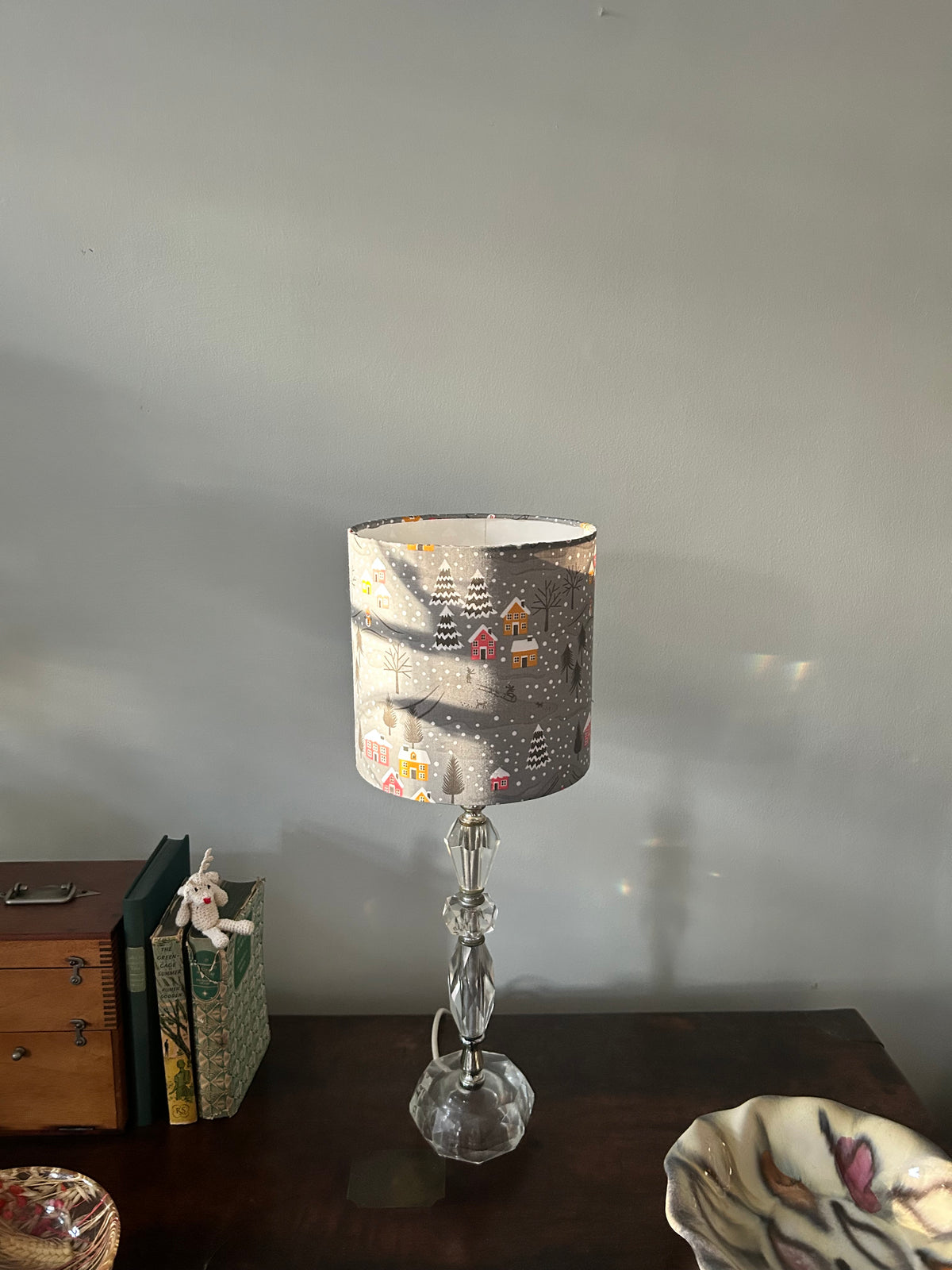 Full view of a glass table lamp with a grey Christmas lampshade, styled with books and a decorative dish.