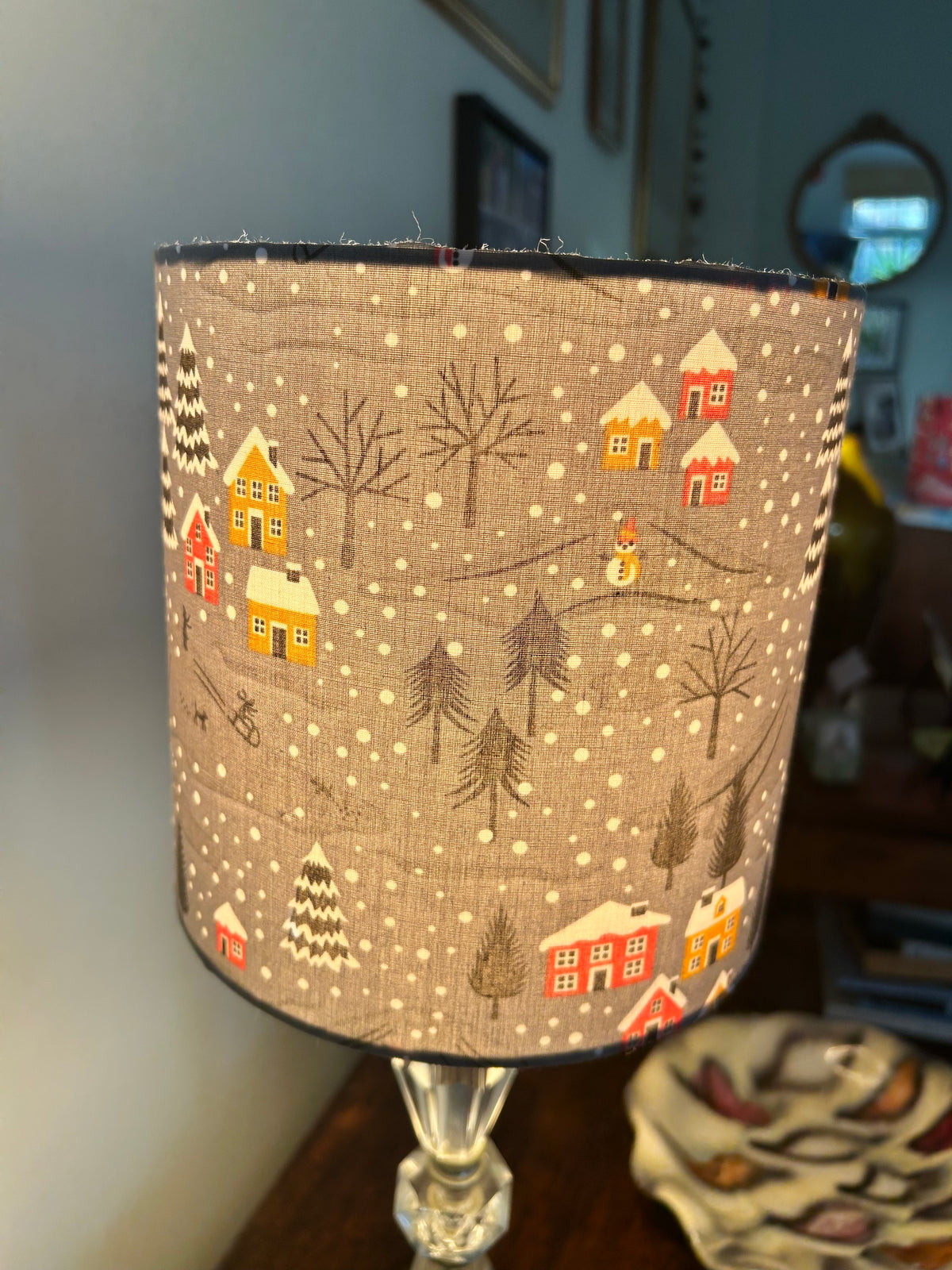 Illuminated grey Christmas lampshade showing its snowy village print glowing warmly from within.