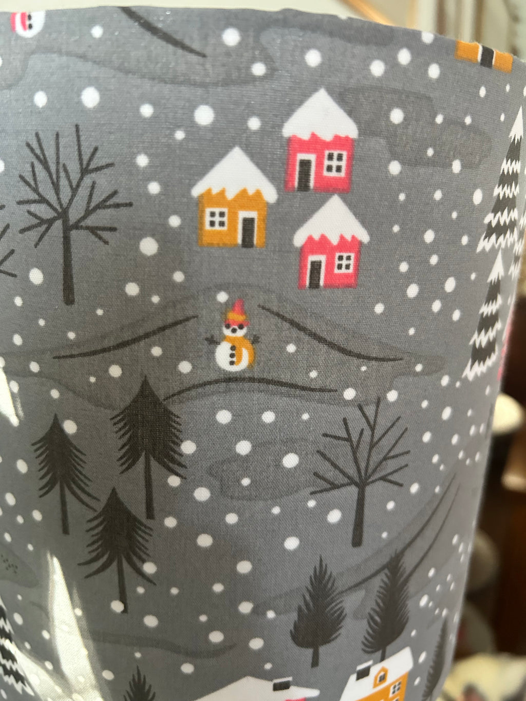 Detail of a snowman beneath raspberry and mustard cottages, surrounded by trees and white snowflakes.