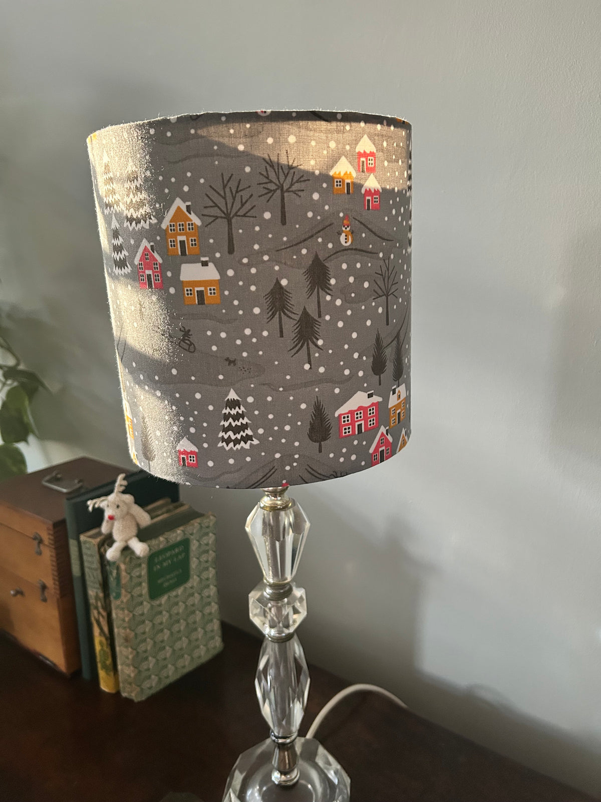 Grey snowy village drum lampshade on a clear glass table lamp beside books on a wooden sideboard.
