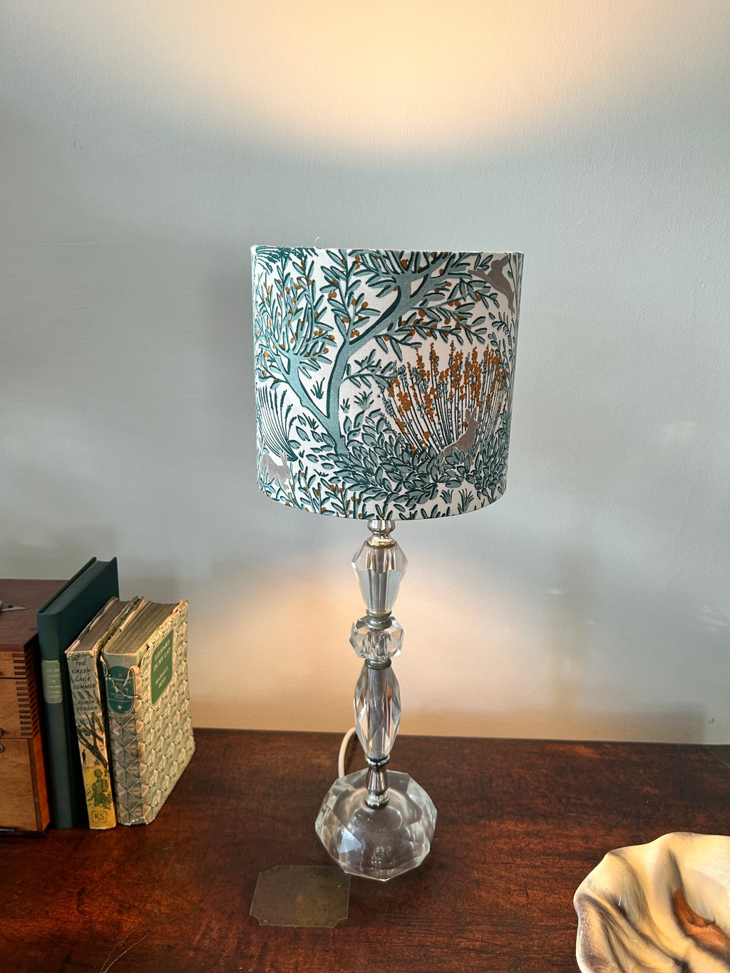 Illuminated woodland rabbit lampshade on a clear glass table lamp beside books on a wooden sideboard.