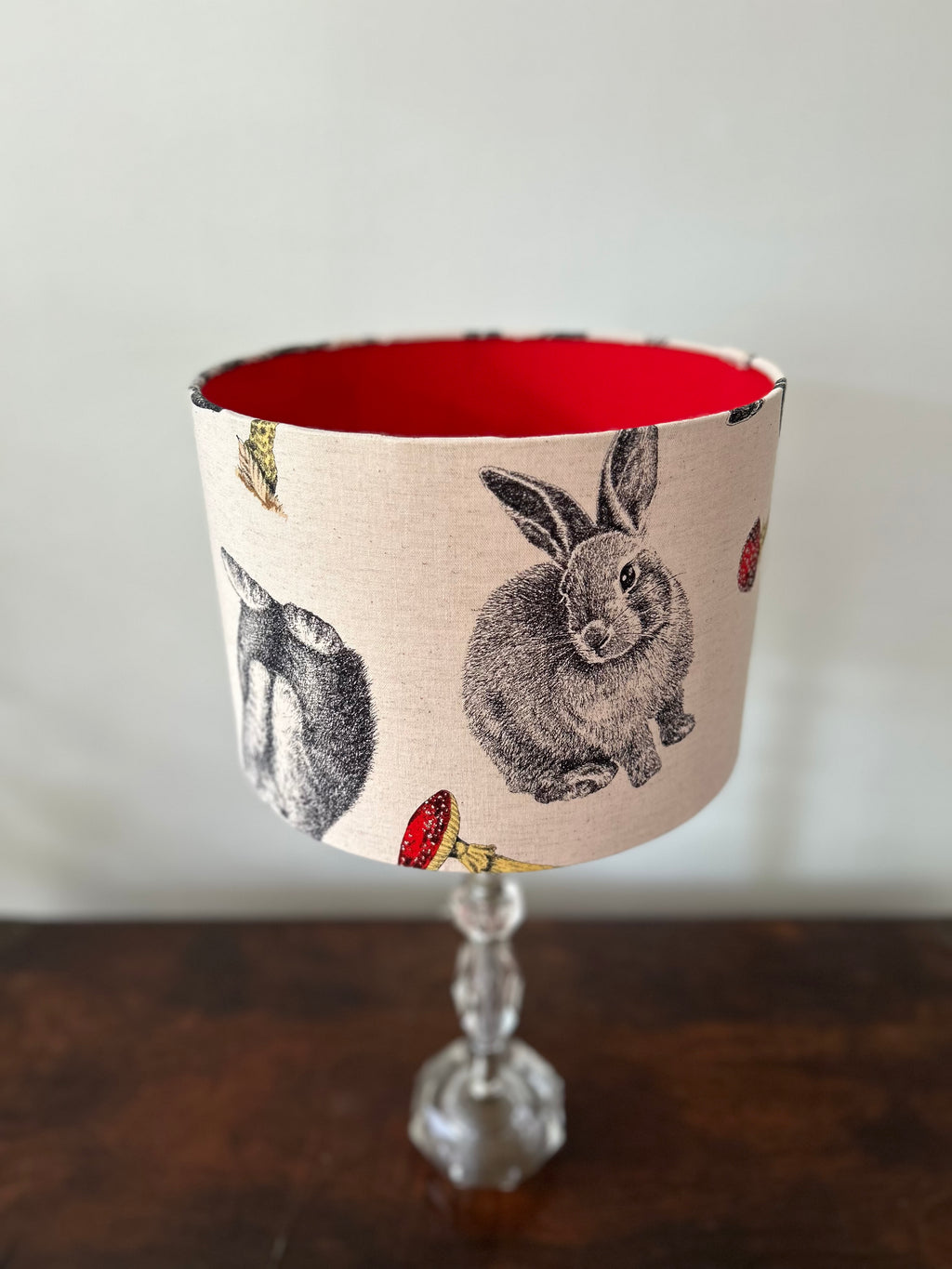 A fabric lampshade with a vintage-style rabbit and toadstool print on a beige background, featuring a bright red interior and a clear glass stem base, set on a dark wooden table against a light gray wall.