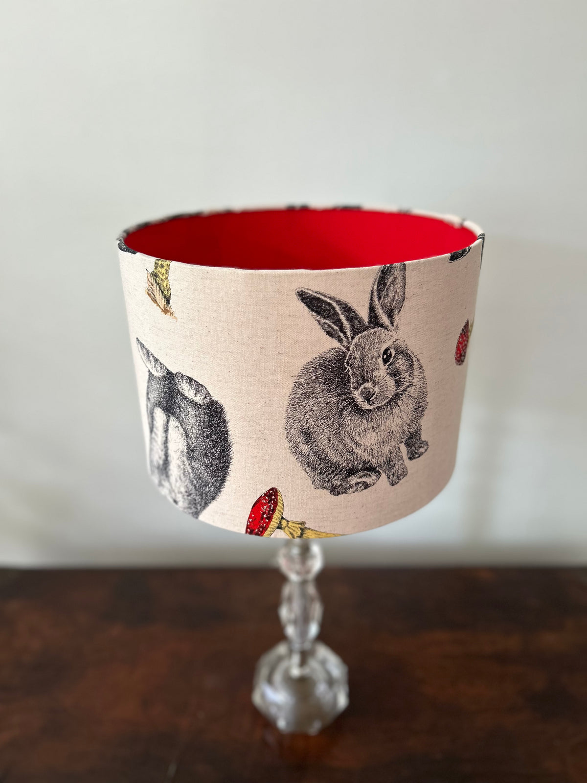 A fabric lampshade with a vintage-style rabbit and toadstool print on a beige background, featuring a bright red interior and a clear glass stem base, set on a dark wooden table against a light gray wall.
