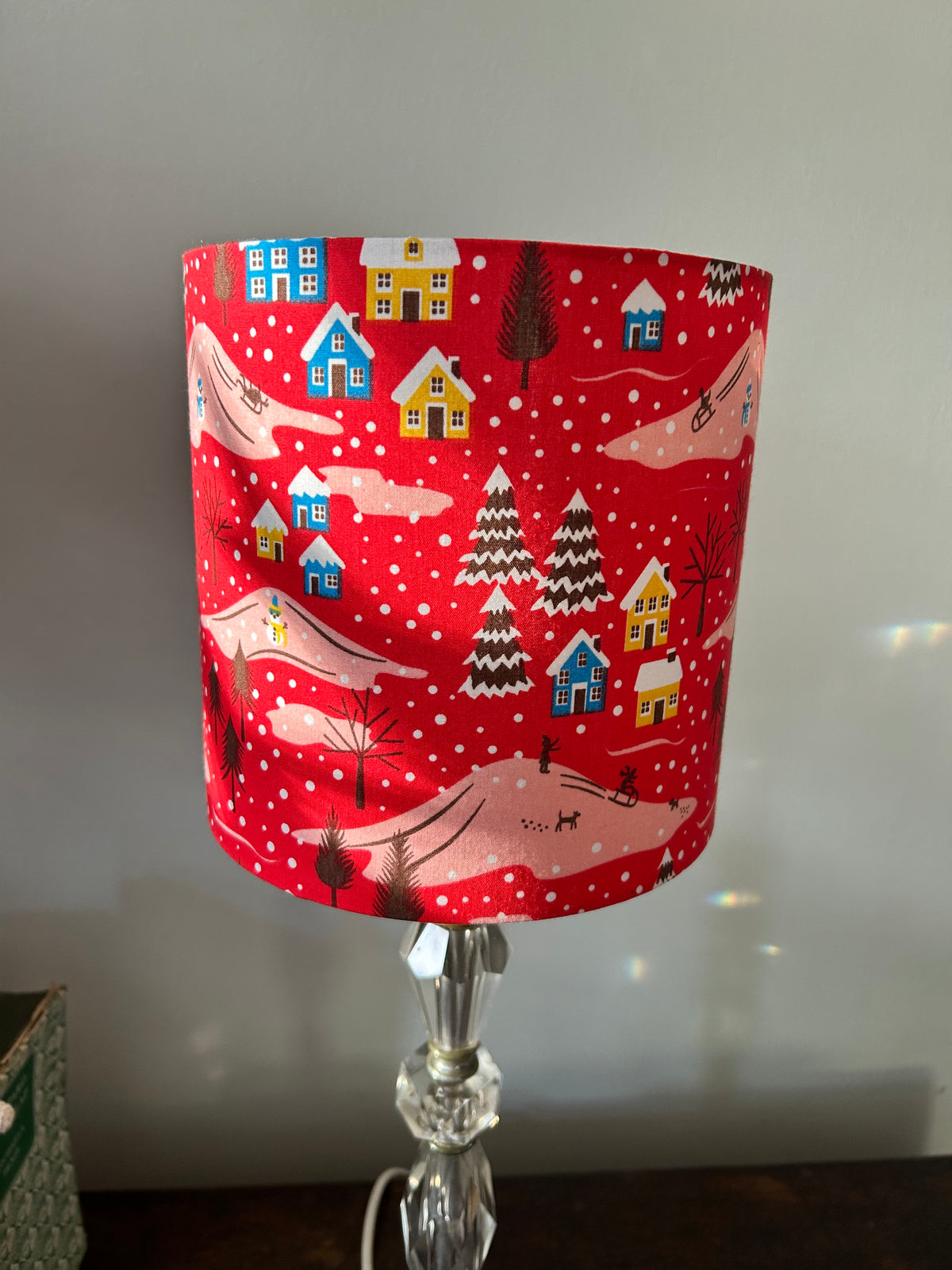 Unlit red Christmas drum lampshade showing its snowy village print on a glass table lamp.