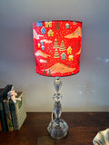 Red snowy village drum lampshade on a clear glass table lamp beside books on a wooden sideboard.