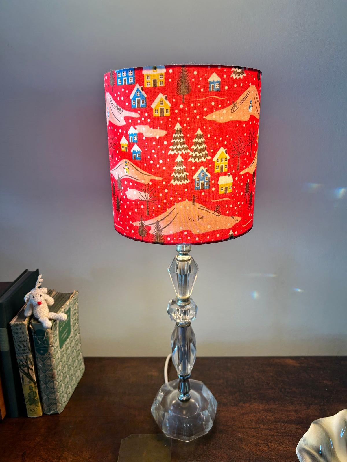 Red snowy village drum lampshade on a clear glass table lamp beside books on a wooden sideboard.