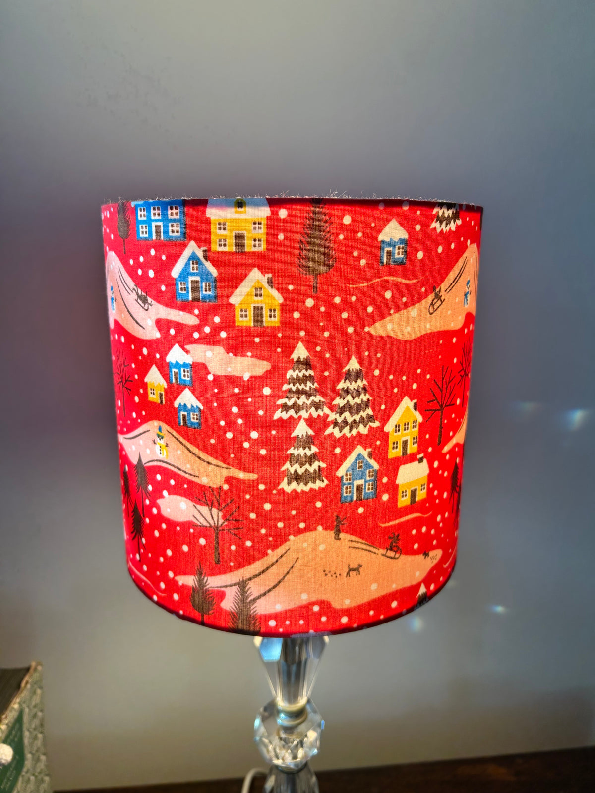 Front view of the red Christmas lampshade with blue and yellow houses, snowy trees and pink hills.